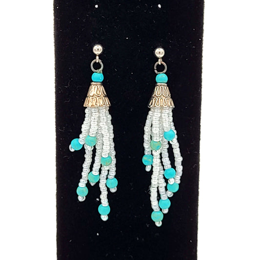  Dangly earrings made of white seed beads with blue beads on the end of each strand. Silver colored end caps and earring hooks on a black velvet earring display.